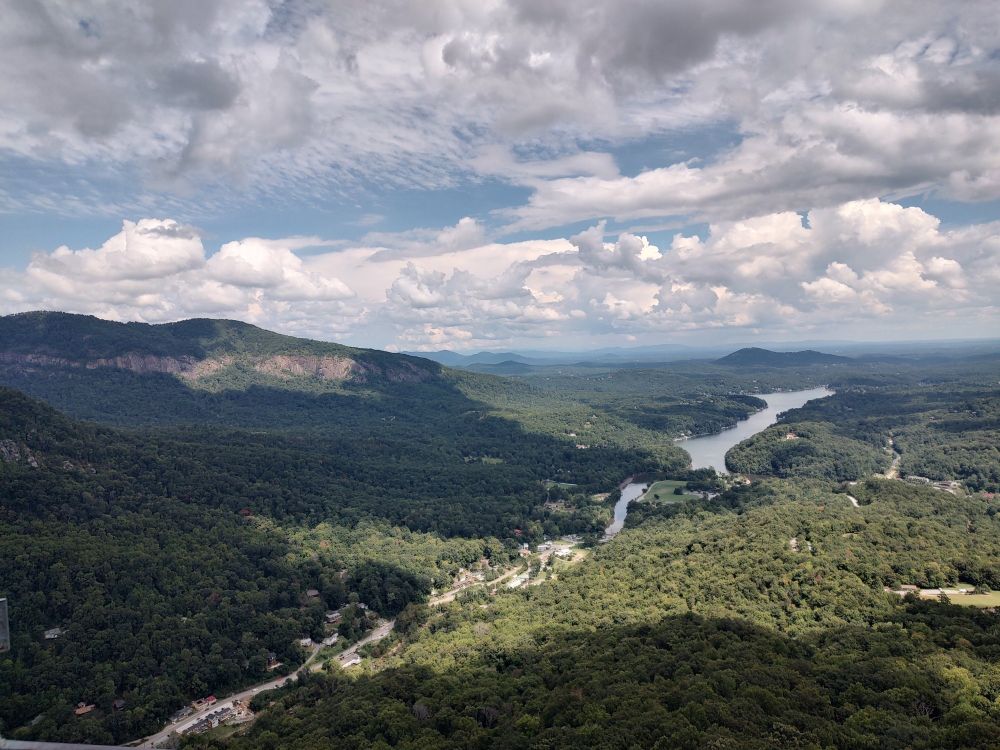Mountain top view out towards Lake Lure, can see green forest covered mountains for miles on end. Closer to you, you can see the stream from the lake starts to fade into the street coming up the mountain where there are many tiny souvenir shops