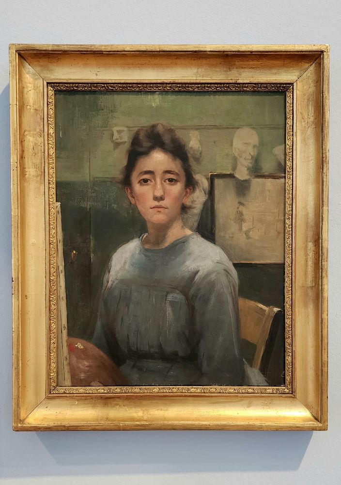 A self-portrait by Augusta Roszmann, 1885-1890, shows a young woman in a blue-grey dress, staring directly at the viewer with an almost neutral expression but also as if she might burst into tears at any moment. Her brown hair is pulled back and she has a painter's palette in one hand