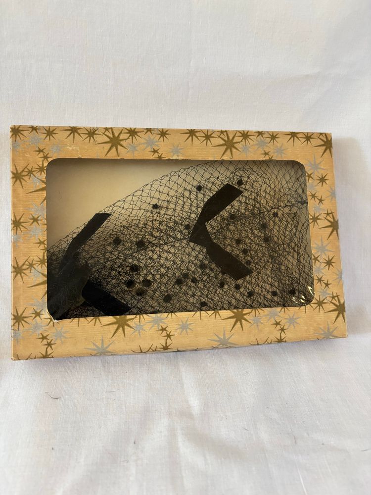 A flat cardboard hat box with star patterns on the box and a black millinery net hat inside. 