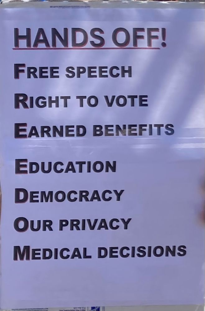 Sign says Hands Off! in black with red outline at top, and below that are these words/phrases:Free Speech, Right to vote, Earned benefits, Education, Democracy, Our privacy, Medical decisions. First letters of each word/phrase below spell “Freedom” on left from top to bottom.