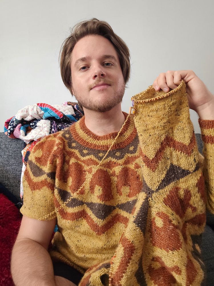 A man is holding up a sweater he is knitting while wearing a very similar one. The yarn he is knitting from is coming from the sweater he is wearing.