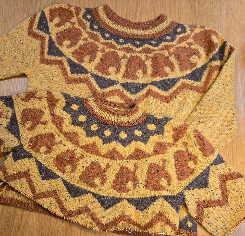 Two sweaters laid out on a wood table. The top is the current design for the Tomnooki sweater and the bottom is the new in-progress redesign. The redesign releases this September!