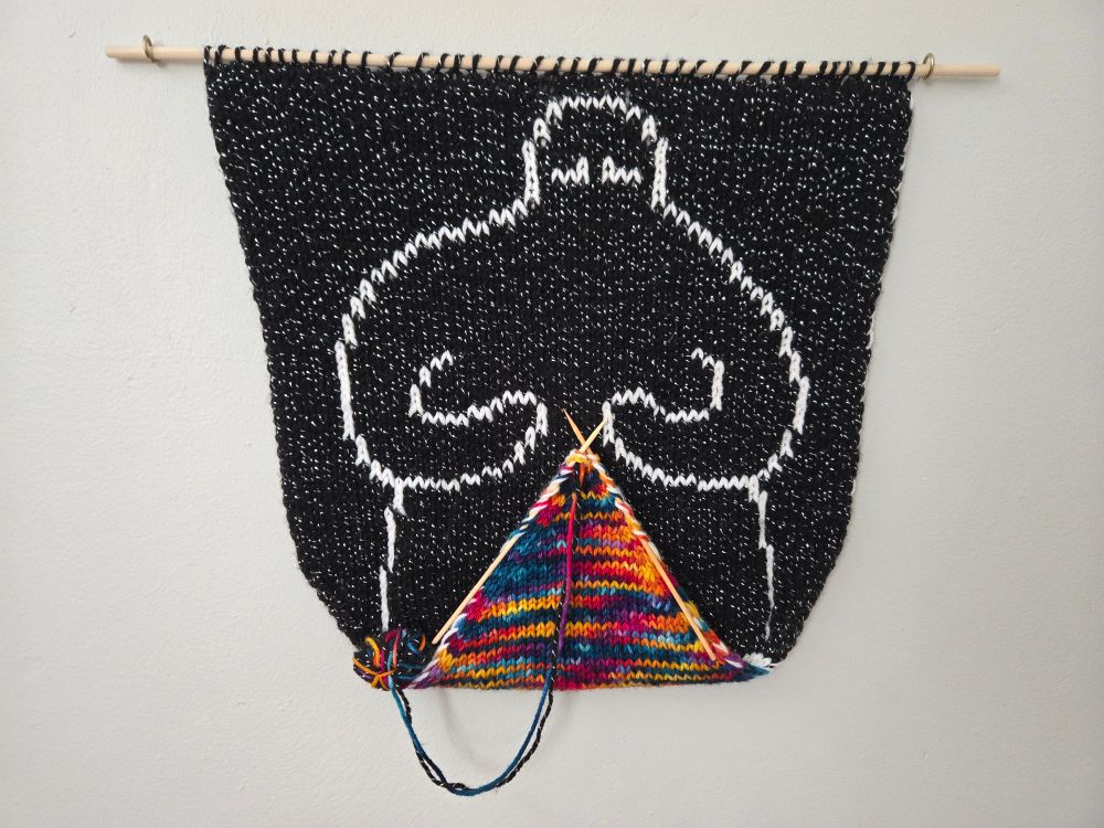 A knitted wall hanging. A figure outlined in white is situated in black and is knitting a triangle in the colors of a sunrise