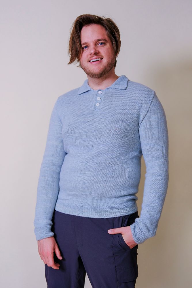 A white man stands in a blue long-sleeved polo with his hands in his pocket