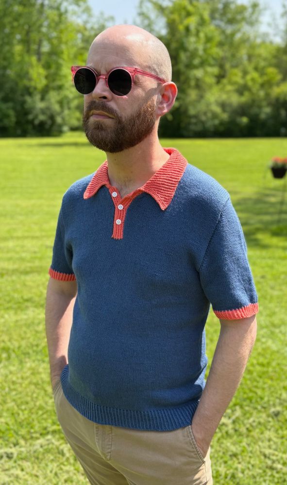 A man stands outside with hands in his pocket wearing a blue and red polo and wearing sunglasses