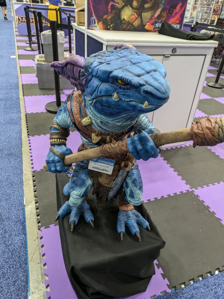 Life sized statue of a short, blue, reptilian humanoid (a kobold). It has people horns, wears a brown leather loincloth and bandolier, and a necklace of bones. It is wielding a spear and has a serious or threatening look on its face.  It is also wearing a GenCon exhibitor badge with the name Maxi Arlo Snarecatcher.
