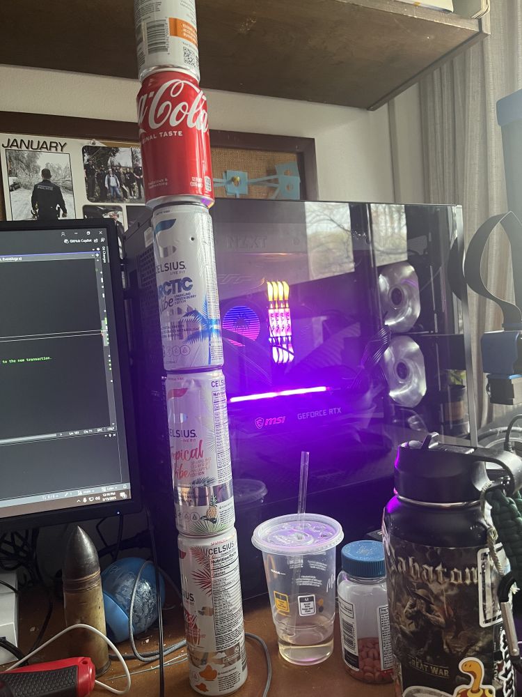 photo of a desk with a stack of Celsius energy drinks on it. other items on the desk include a PC, a bottle of advil, a water bottle, an empty mcdonalds coke, an electric screwdriver, and an anti-tank round (disabled).