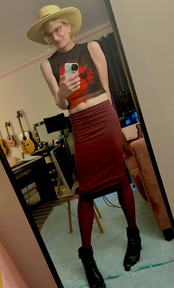 Angela, a middle aged blonde trans woman, takes a mirror selfie wearing a wide brimmed straw hat, a sleeveless black top and a maroon tube skirt. 
