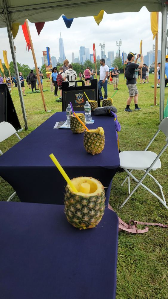 5 pineapples with straws sit on two blue staff tables under a tent. The GO Fest battle area event is going on the background with multiple people. You can see the city in the very background.