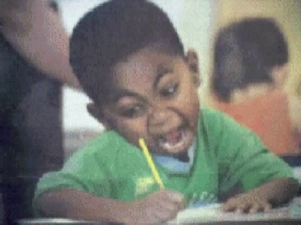 a young boy in a green shirt is writing on a piece of paper with a yellow pencil in his mouth .