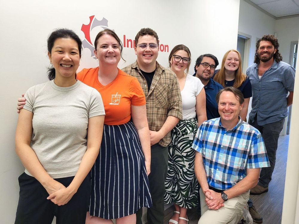 Kristina, Courtney, Tyler, Shanelle, Jake, Justin, Josie, and Reed standing in front of the Innovation gear logo in our Burnaby office.