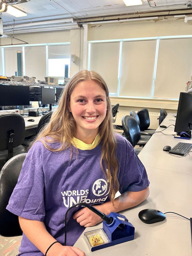 Clubs That Care youth leader Skye wearing a Worlds UNBound shirt and sitting in front of a soldering iron.