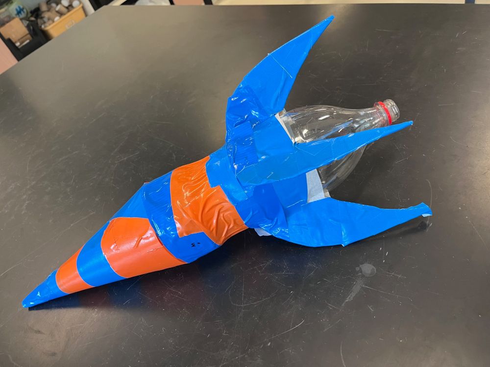 Student-made rocket with cardboard fins attached to 2 liter bottle/rocket decorated with blue and orange duct tape