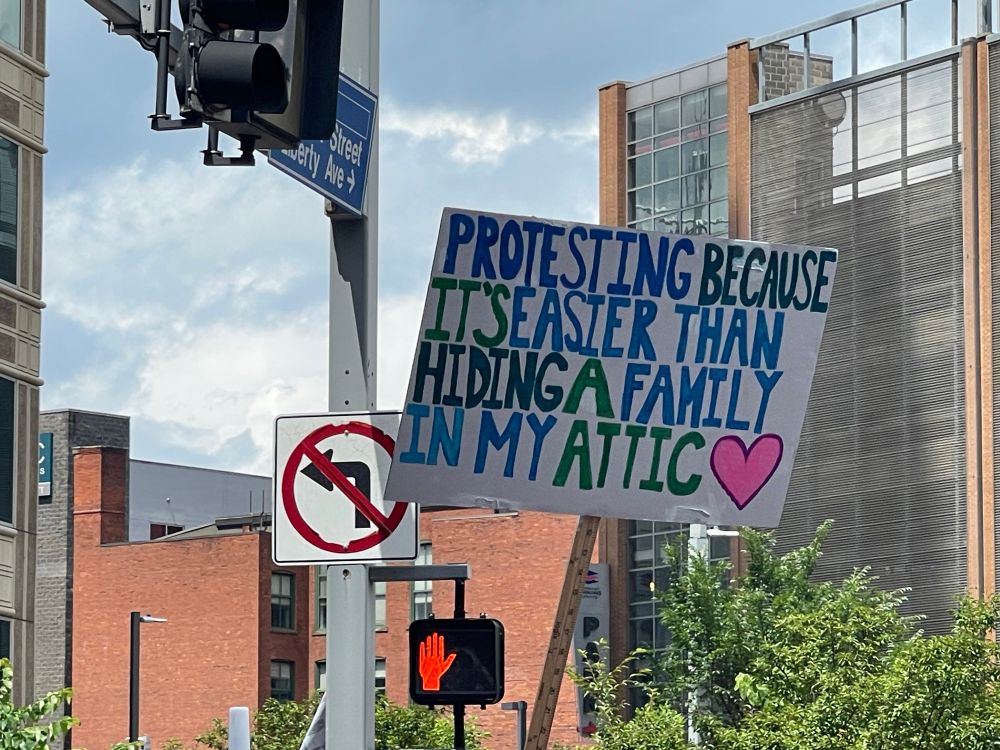 Protest sign on white postcard that reads: “Protesting because it’s easier than hiding a family in my attic”. 