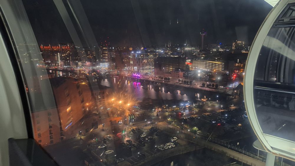 A view of Liverpool docks at night,  from up high,  taken from within a big wheel pod. There are multiple glows from different types of city light. 