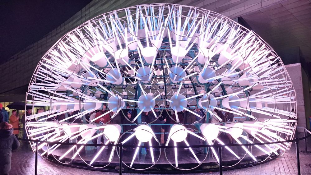 A semi circle sculpture of light,  made from cones and tubes, glowing pink and white.  Like a light hedgehog. 