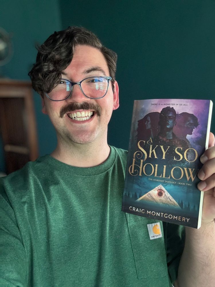 Craig, a white man with dark hair, is in a green shirt and is holding a copy of A Sky So Hollow