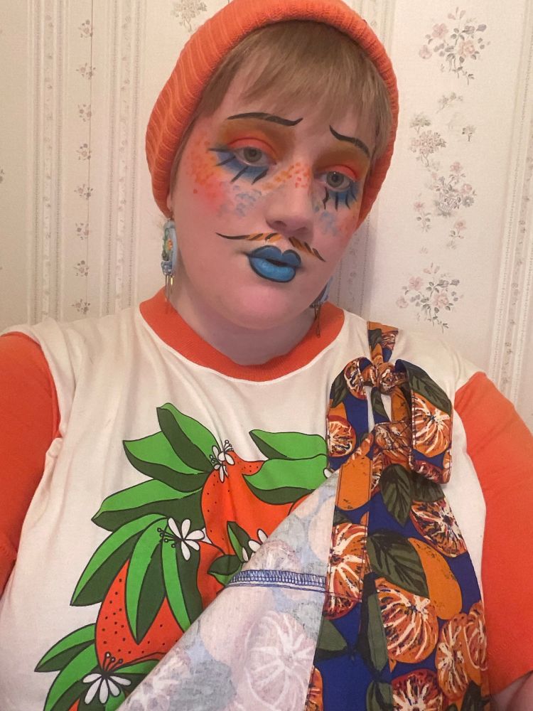 White nonbinary person with blonde hair wearing an orange printed graphic tee with orange printed overalls and an orange beanie. 