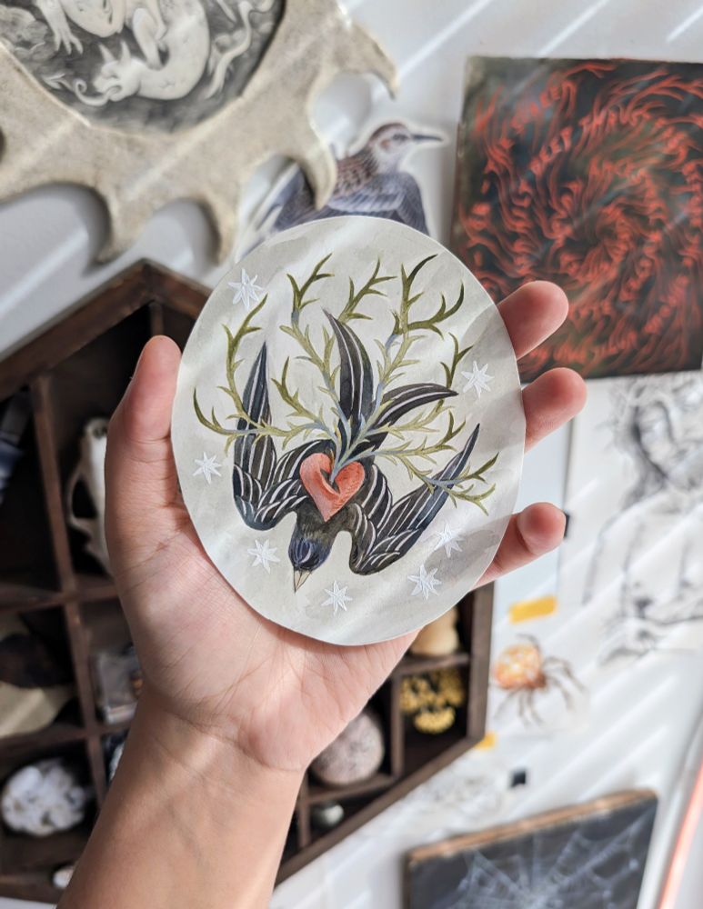 A hand holding an oval painting. The painting is of a dark bird diving with a red heart in the center. The heart has green thorns coming out of it. The background is light grey with white stars. In the background is a wall with various art pieces hanging. There are light beams shining. 