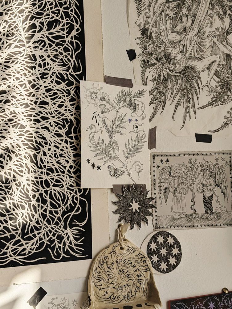 An art wall with various black, white and grey drawings. These depict a vetch flower, a sun and moon with stars, a demon and angel holding a sun and moon in a medieval style, a replication of St Anthony being tormented by demons, and a root system with a black background. Below is a ceramic shelf with spiral metal writing. 