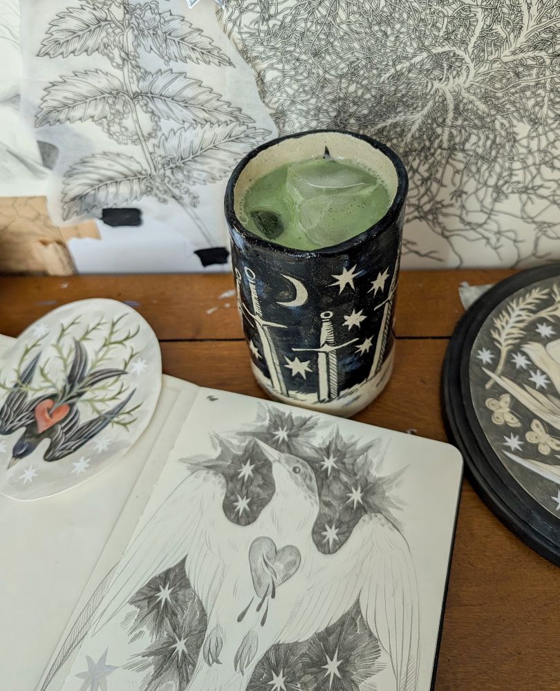 A cup with iced matcha in it, on a desk with a sketchbook and painting. The painting is of a bird diving with a thorny heart in the center. The sketchbook is of a tern with a bleeding heart in the center. The wall has botanical drawings on it. The cup is handmade ceramic with a sword star and moon pattern on it with a black background. 