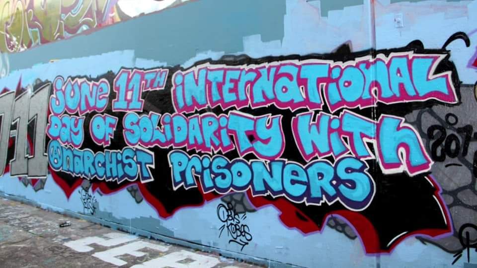 A photo of a graffitied wall that reads "June 11th International Day of Solidarity with Anarchist Prisoners" in blue bubble letters.