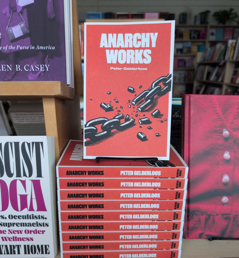 A photo of a stack of books, "Anarchy Works" by Peter Gelderloos, inside Firestorm Books. The books are flanked by other titles, including "Fascist Yoga" by Stewart Home and  "The Handsewn Wardrobe" by Louisa Owen Sonstroem.