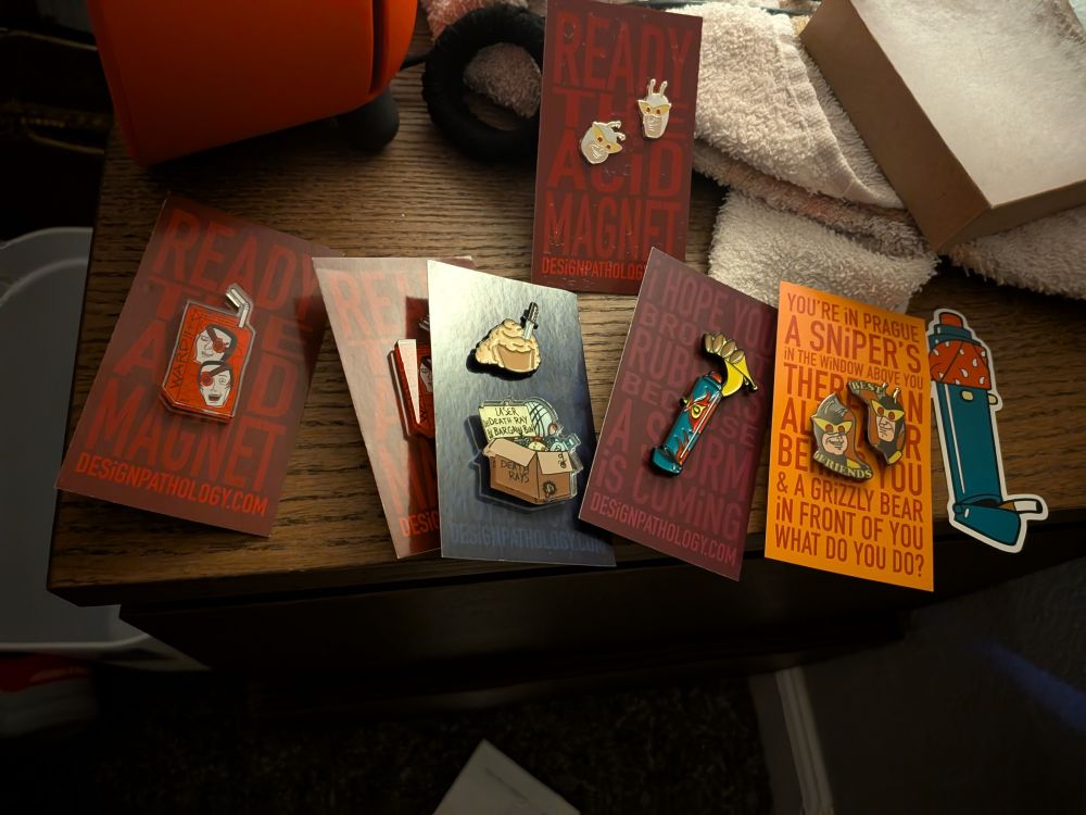 A photo of venture bros merch, from left to right: 
A pin of a juice box labeled ward with the heads of watch and Ward on the box, a pin of a cupcake with a blonde curly mullet with a knife sticking out, a pin of a cardboard box labeled laser death ray bargain bin, a pin of car paint job HELPeR from the episode dia de los dangerous, best friends pins of henchmen 21 and 24, a sticker of helper wearing a bandana and smoking a cigarette, and a pair of earrings of the faces of 21 and 24 
