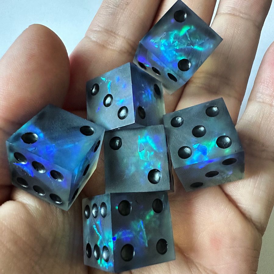 A handful of six pip d6 dice. They are dark gray with blue and green mylar inside, creating a glowing effect.