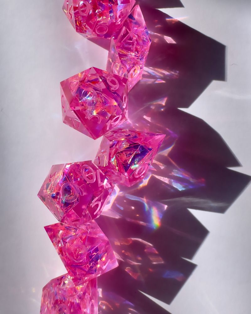 A set of pink holographic dice in a vertical line. The sunlight on the dice casts prismatic reflections on the white backdrop.