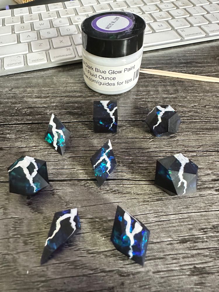 A set of unfinished dice blanks. The dice are black with blue/green mylar inside, and there are white lightning bolts painted on the dice. There is a small bottle of blue glow in the dark paint in the background.