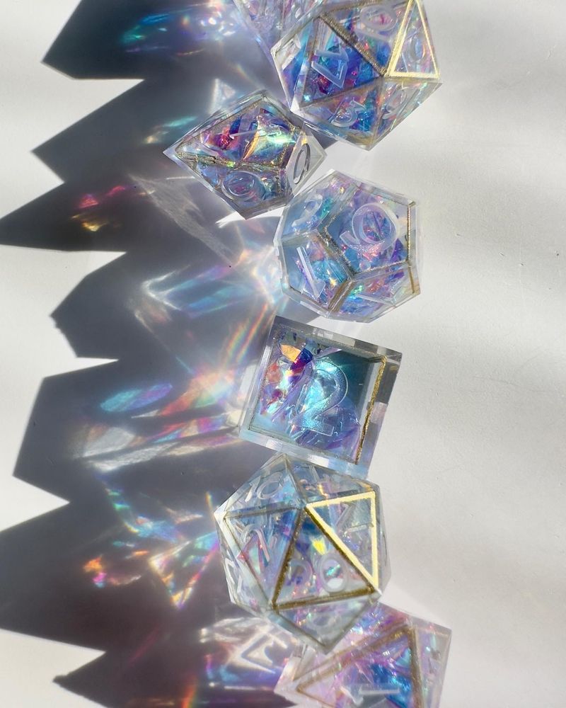 An unfinished pastel dice set with holographic mylar, lined in gold. The sunlight on the dice cast rainbow patterns onto the white background.