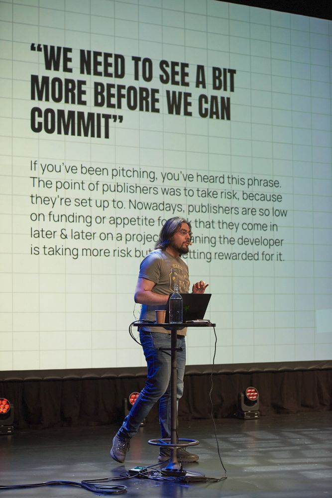 Rami speaking on a stage. The slide describes the lack of risk-taking by publishers.