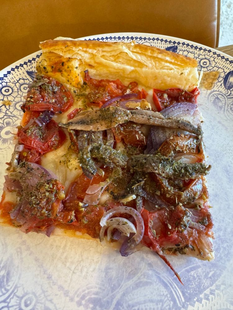 A slice of riganada on a plate with a faded blue pattern. 

Puff pastry with sliced tomatoes, onions, anchovies and A LOT of oregano baked in the oven. 


