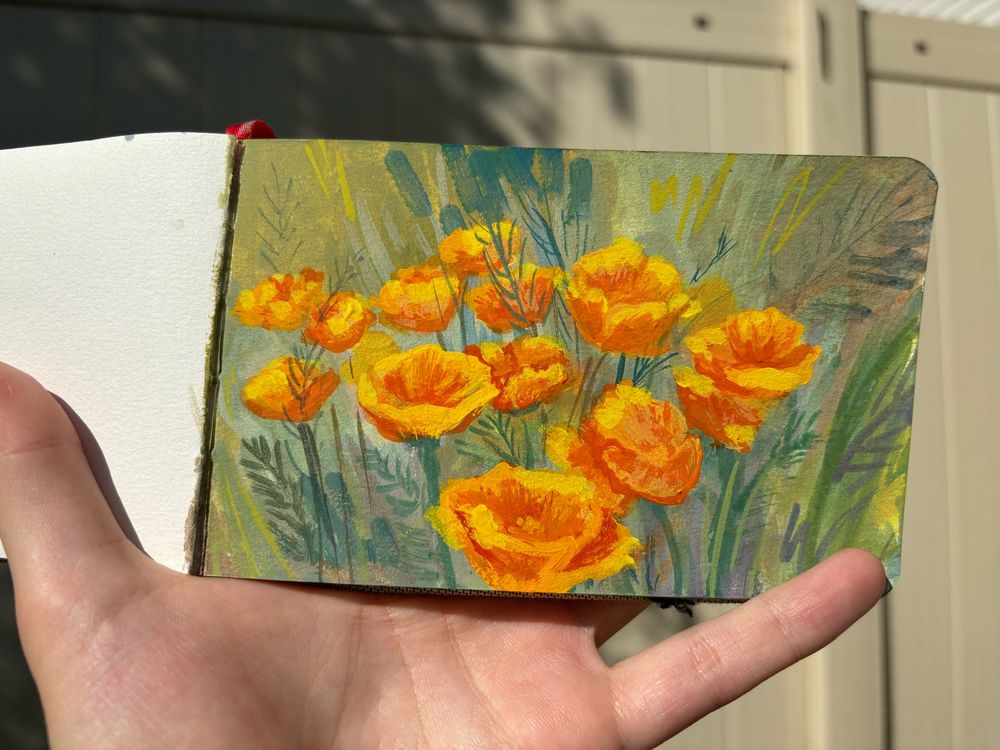 Hand holding a small sketchbook with a painting of bright orange poppy flowers. The background has multiple shades of green and leaves painted quickly around. The flowers have more details than the rest of the piece, but the whole painting has a “painterly” style to it. 