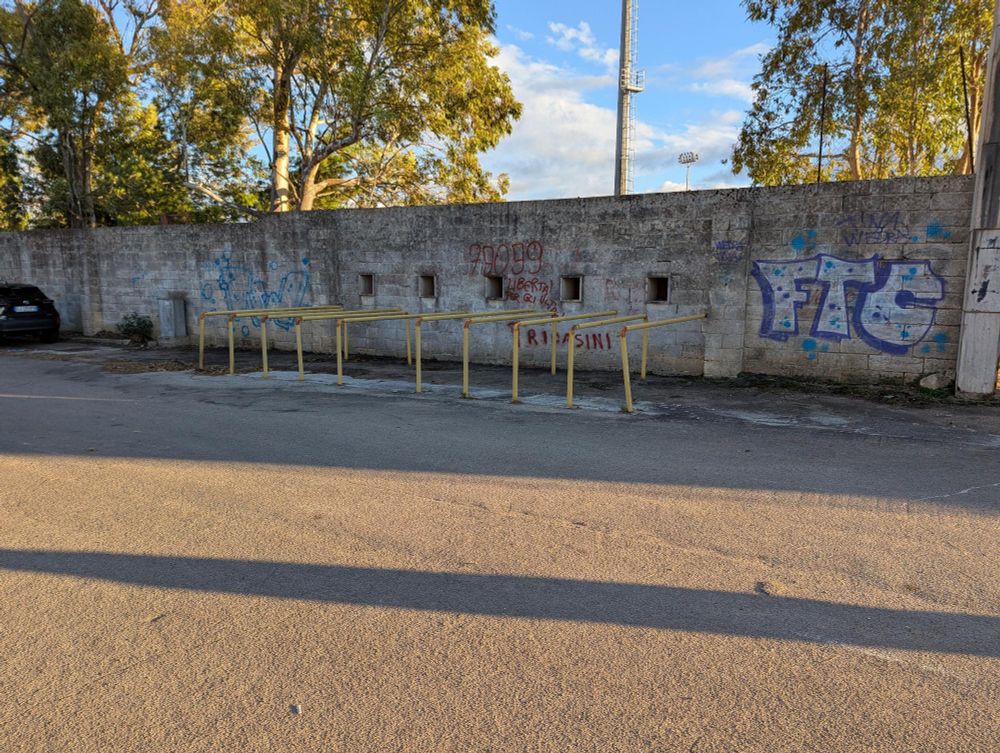 Blick über einen Platz auf alte Ausgabefenster von Tickets mit Absperrbarrieren. Hinter der mit Grafitti besprühten Mauer stehen Bäume, links im Bild parkt ein schwarzes Auto. 