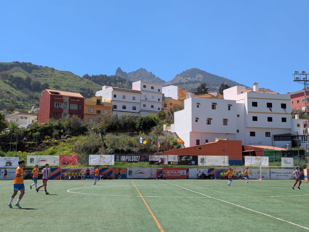 Blick über ein Fußballfeld. Links und rechts im Bild sind Spieler zu sehen. Im Hintergrund stehen Wohnhäuser und dahinter sind Berge zu sehen. 