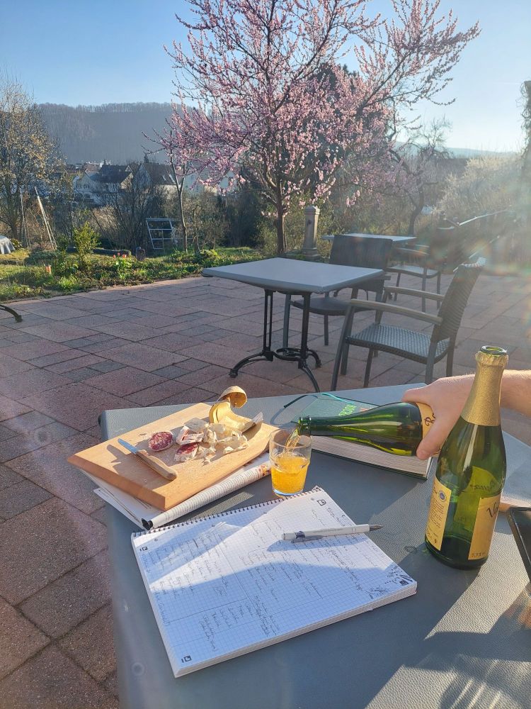 Tisch auf Terrasse in der Sonne, blühender Pfirsichbaum im Hintergrund, Cidre und Salami auf dem Tisch neben handschriftlichen Notizen