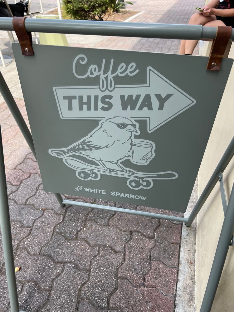 Coffee shop sign that says “Coffee this way” with an arrow pointing to the shop and a sparrow wearing sunglasses on a skateboard holding a cup of coffee