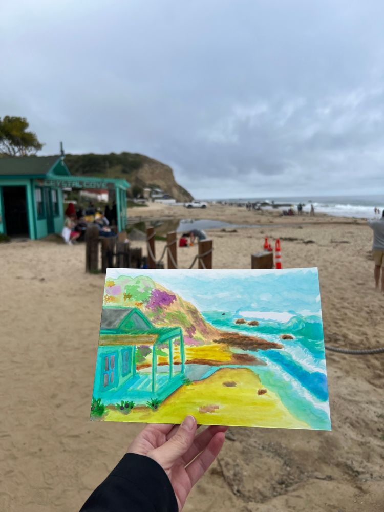 Watercolor painting of cottage and beach shoreline held in front of the painted landscape