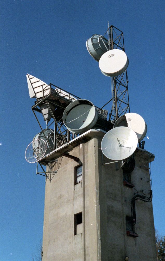a square concrete building with various dish and horn shaped antennas crowded on the roof against a clear blue sky