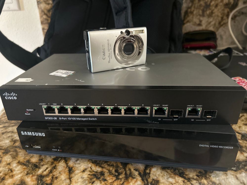 A black Samsung DVR is on top of a yellowed granite table, on top of it there's a cisco branded network switch, on top of it theres a canon coolpix