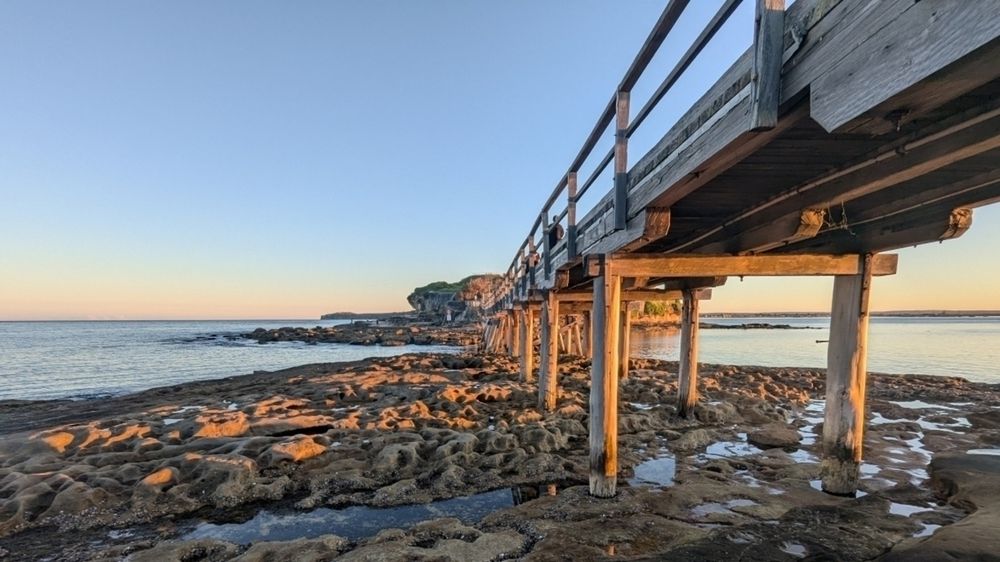 A wooden pier extends over a rocky shoreline at sunset, with a calm body of water and a distant landmass in the background.