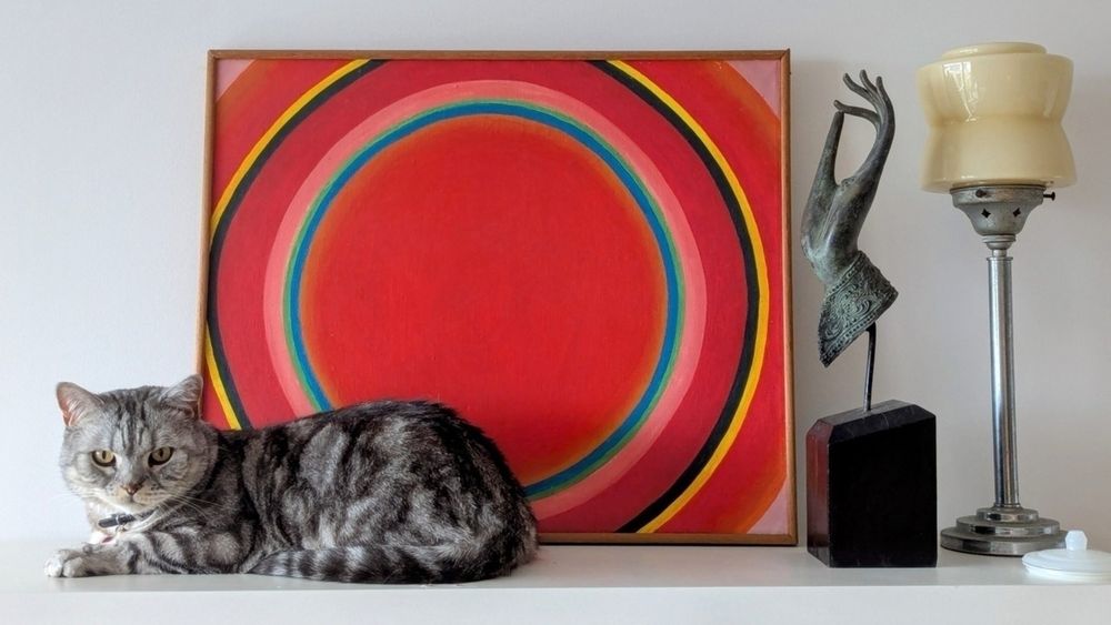 A striped gray cat lies on a shelf next to a vibrant, circular abstract painting, a sculptural art piece, and a vintage lamp.