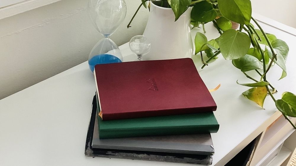 A stack of four notebooks is placed on a white surface next to a potted plant and an hourglass.