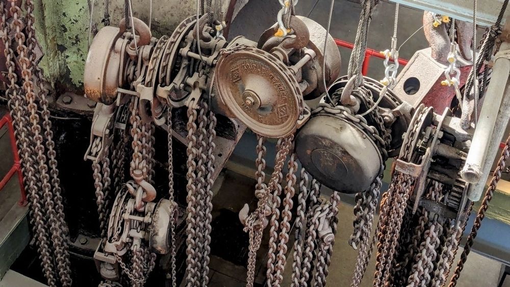 Several hoists with chains are hanging in a workshop setting, featuring a variety of metal components.