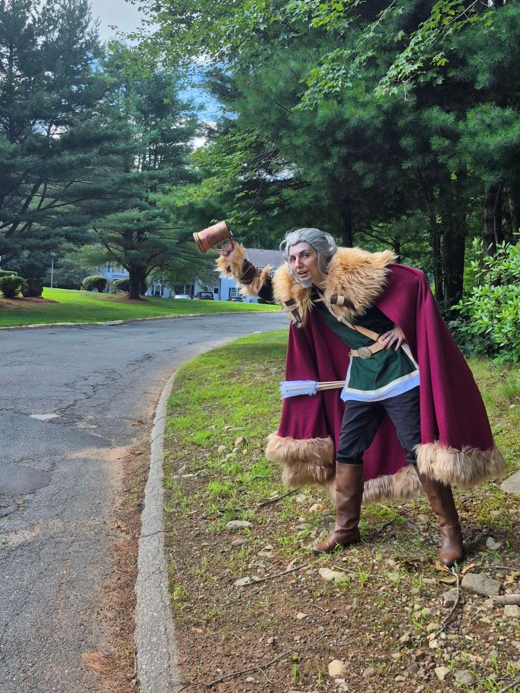 Photograph of a Z’aanta (Octopath Traveler) cosplayer motioning like they are waving down a car with a brass tankard in their right hand.