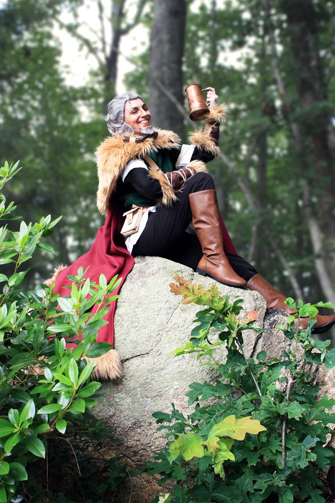 Photograph of a cosplayer sitting on a well lit rock amongst foliage. The cosplayer is in the garments for Z’aanta the hunter from the video game Octopath Traveler. The cosplayer is resting their right forearm atop their raised right knee, and their left arm is raised, holding a brass metal tankard in a cheering motion.
