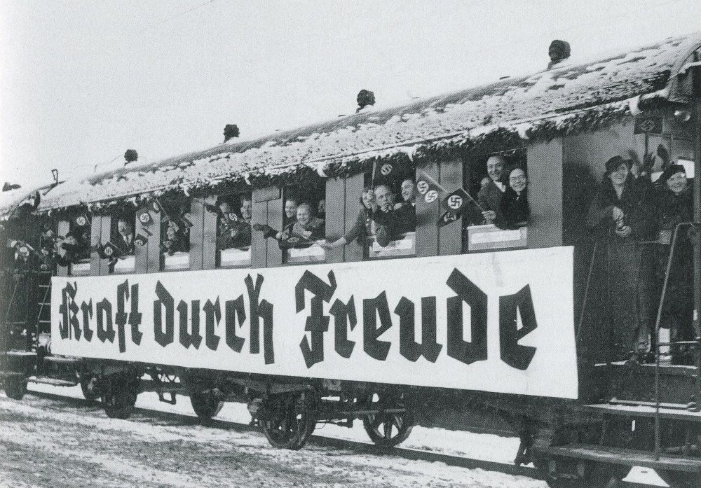 A train car with the slogan "Kraft durch Freude" in large letters, and people hanging out the windows waving small Nazi flags.