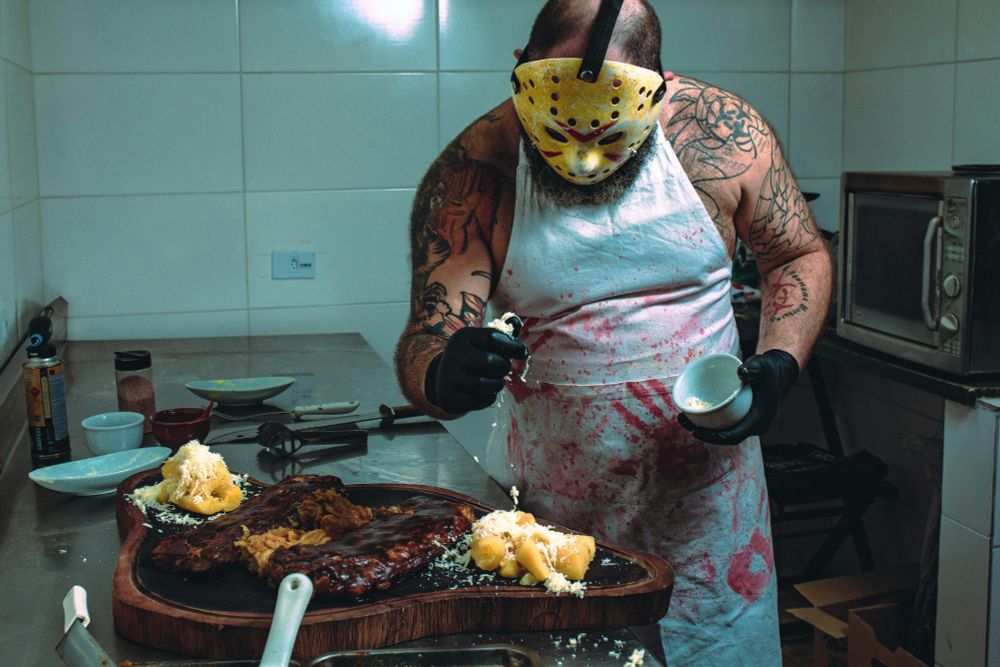 Man in bloody apron and hockey mask in a kitchen salting a wooden platter of ribs and mashed potatoes. 